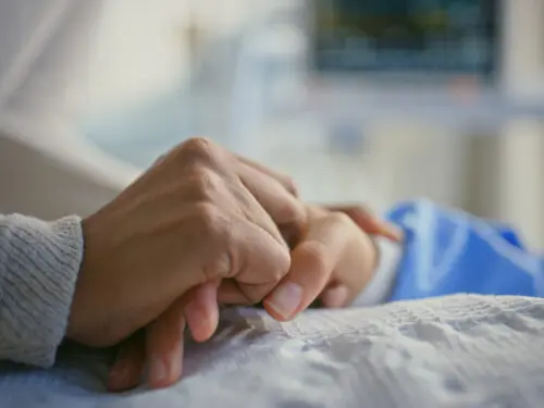 Person holding a patient's hand by hospital bed for support.
